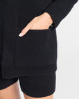Close-up of a person wearing a black ribbed knit sweater with a large front patch pocket and matching black knit shorts, with one hand tucked into the pocket against a light gray background.