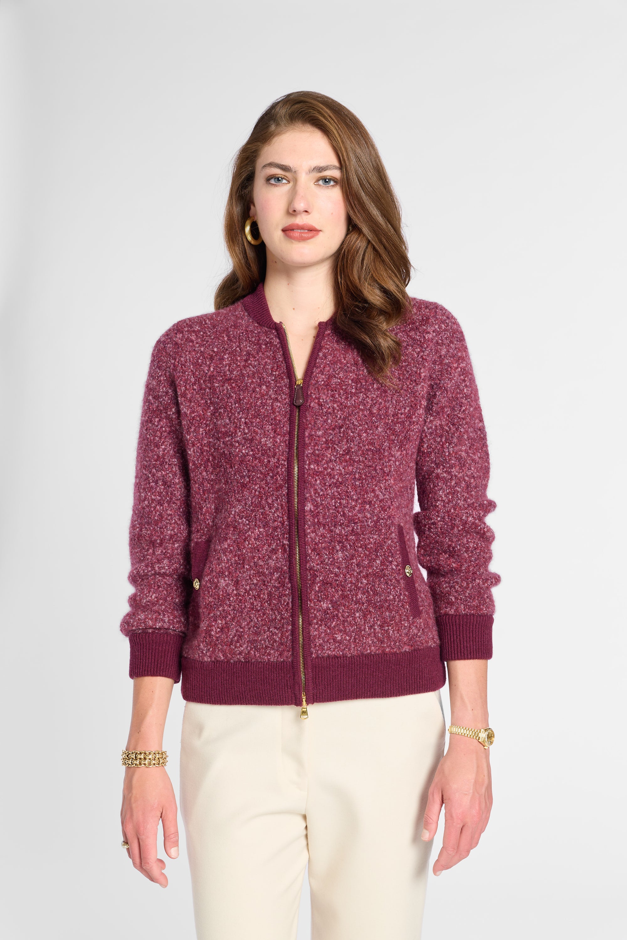A woman modeling a marled burgundy zip-up knit cardigan with ribbed collar, cuffs, and hem, paired with cream pants, against a light gray studio background.