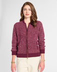 A woman modeling a marled burgundy zip-up knit cardigan with ribbed collar, cuffs, and hem, paired with cream pants, against a light gray studio background.