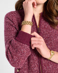 A close-up of a woman wearing a marled burgundy zip-up cardigan sweater with gold-tone hardware, accessorized with gold bracelets and a watch.