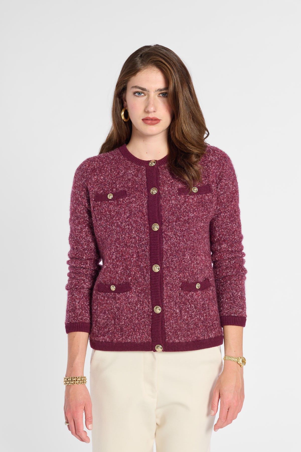 A woman modeling a textured tweed cardigan in burgundy with gold-tone buttons and flap pockets, paired with cream trousers, against a light gray studio background.