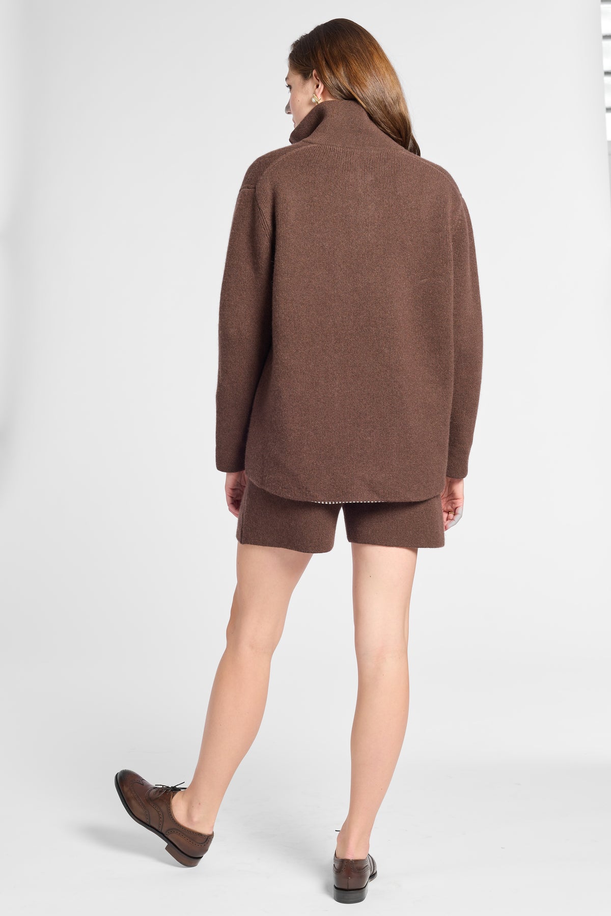 A woman shown from the back wearing a brown knit quarter zip sweater and matching brown shorts, paired with brown shoes, against a light gray studio background.