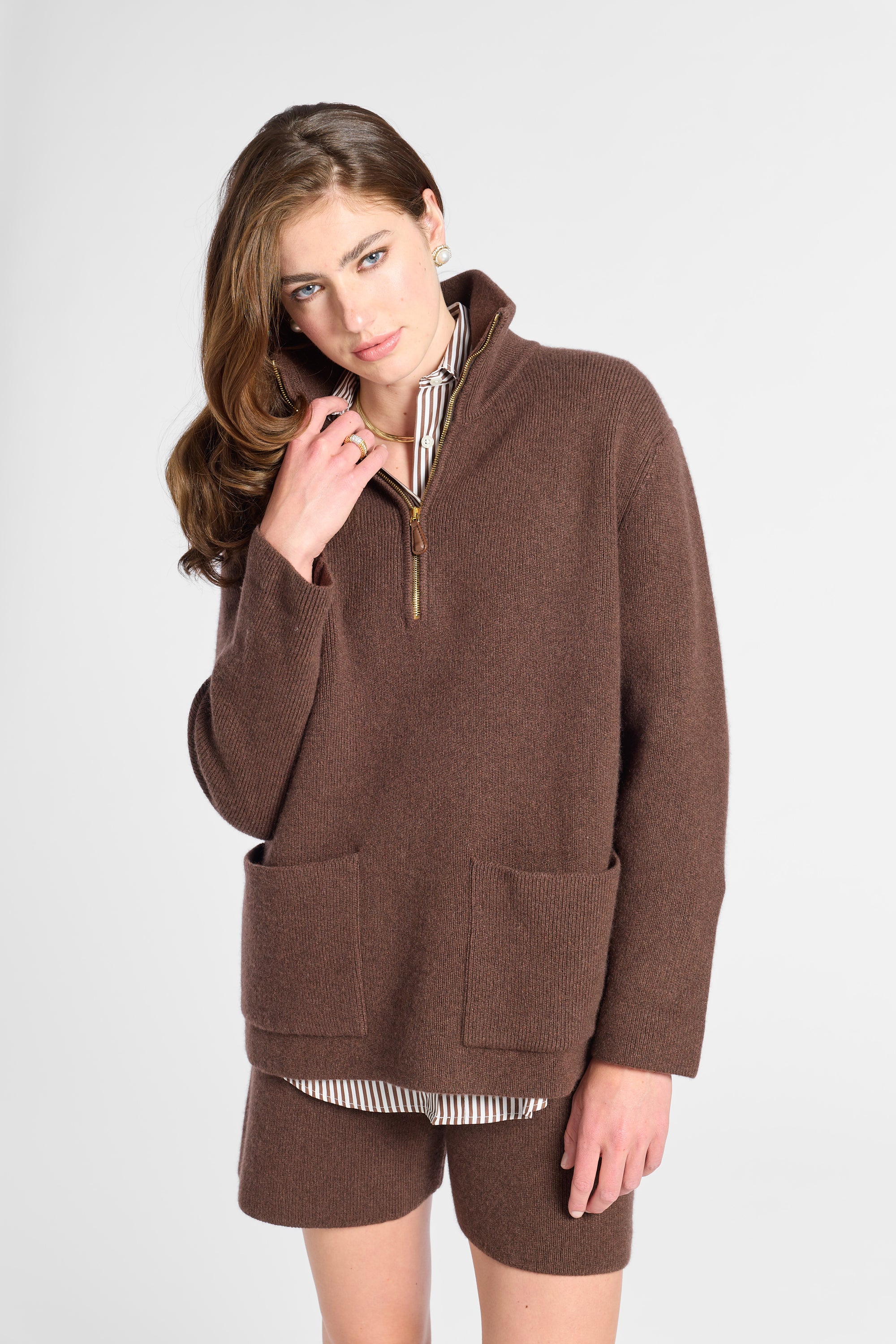 A woman modeling a brown quarter-zip fleece pullover sweater with front patch pockets, paired with matching brown shorts and a striped shirt underneath, against a light gray studio background.