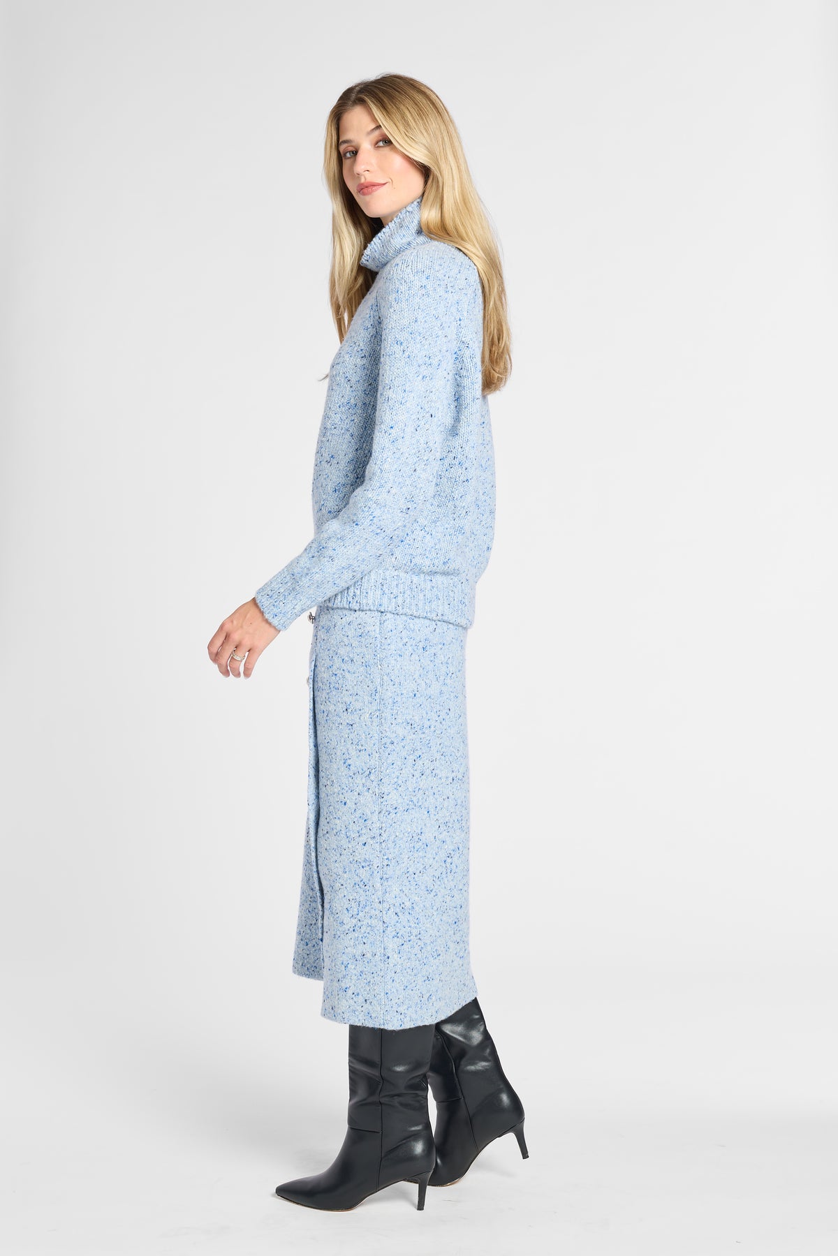 A woman modeling a light blue knit cardigan and matching midi skirt set with a subtle speckled pattern, paired with black heeled ankle boots, against a plain light gray studio background.