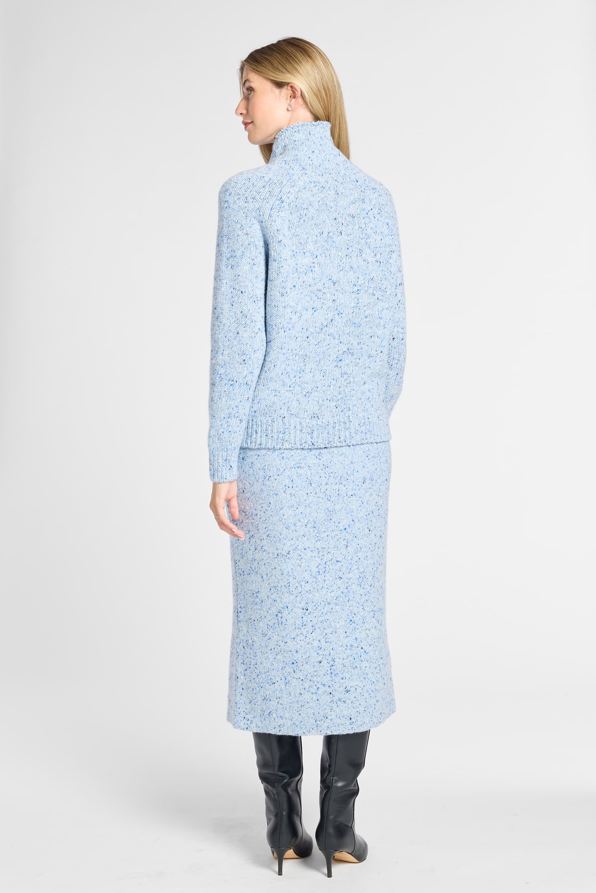 A model shown from the back wearing a light blue knit turtleneck sweater and matching midi skirt set with a subtle speckled texture, paired with black knee-high boots, against a light gray studio background.