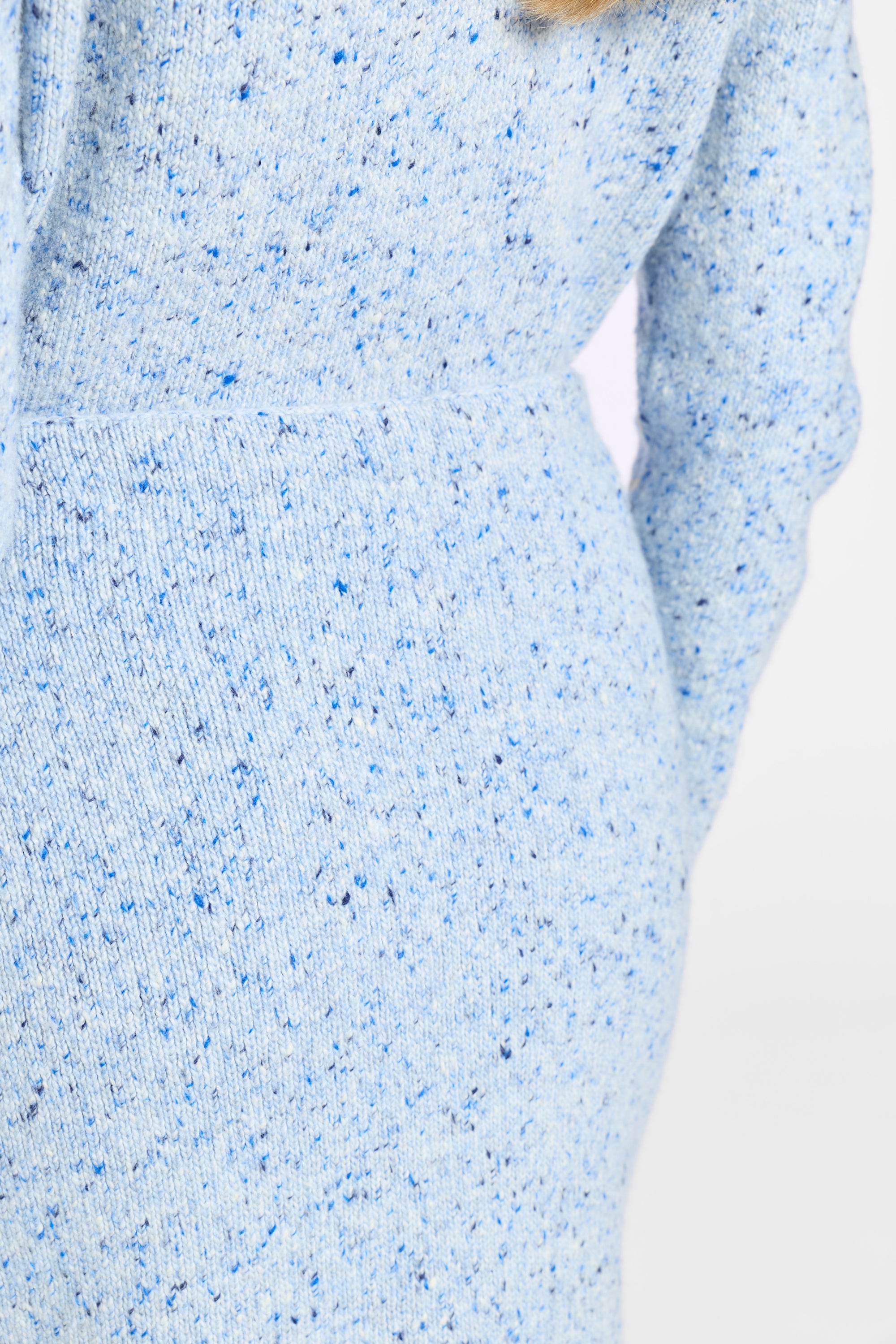Close-up of a light blue knit sweater with blue and black speckles, showing the ribbed texture and part of the sleeve against a plain white background.
