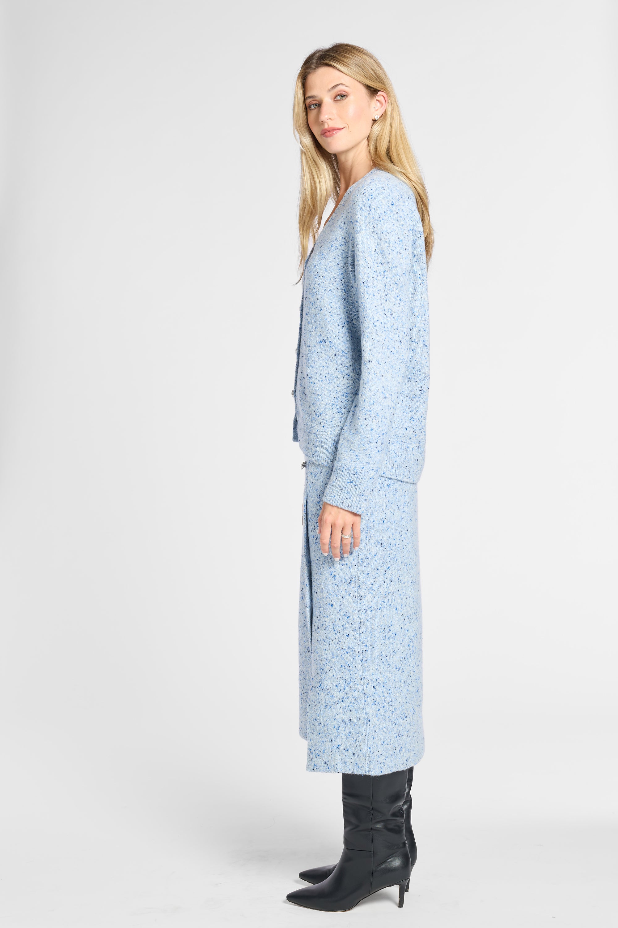 A woman modeling a light blue knit cardigan with a subtle white speckled pattern, paired with black pointed-toe ankle boots, against a plain light gray studio background.