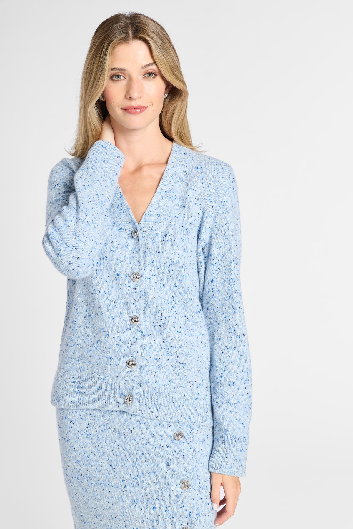 A woman modeling a light blue knit cardigan and matching skirt set with a subtle speckled texture and silver buttons, photographed against a plain light gray studio background.