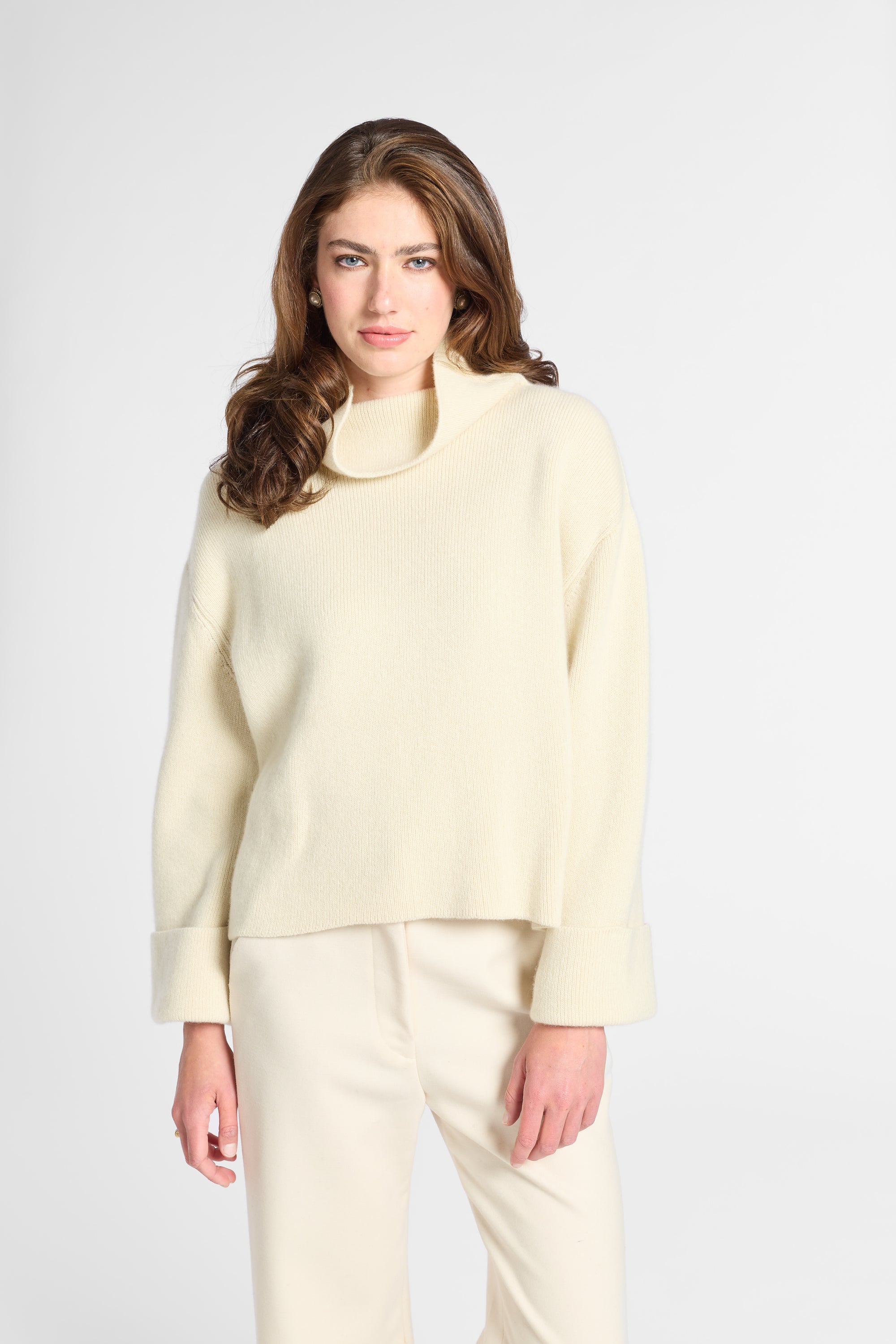 A woman modeling a cream-colored knit mock neck sweater and matching cream trousers against a light gray studio background.