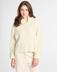 A woman modeling a cream-colored knit mock neck sweater and matching cream trousers against a light gray studio background.