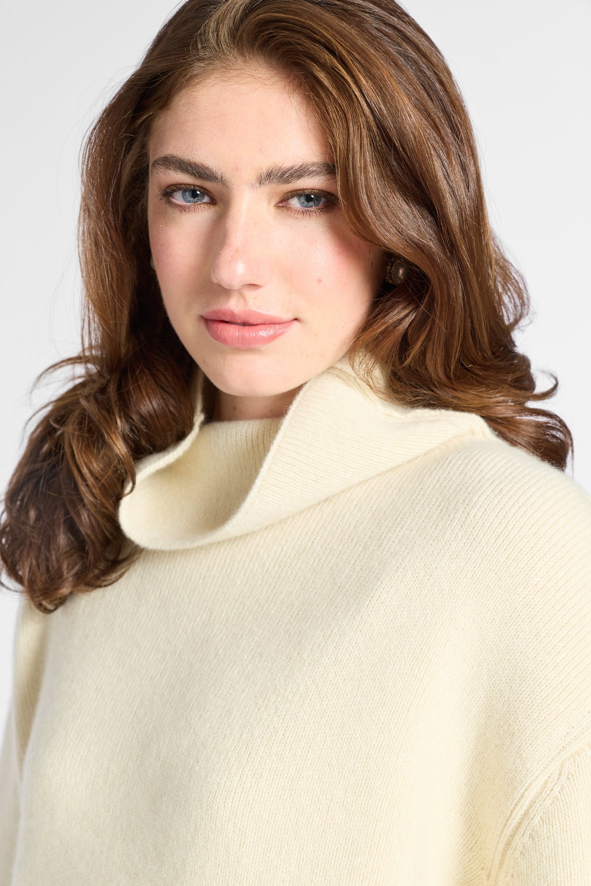 A close-up of a woman with brown hair wearing a cream-colored knit turtleneck sweater against a light gray background.