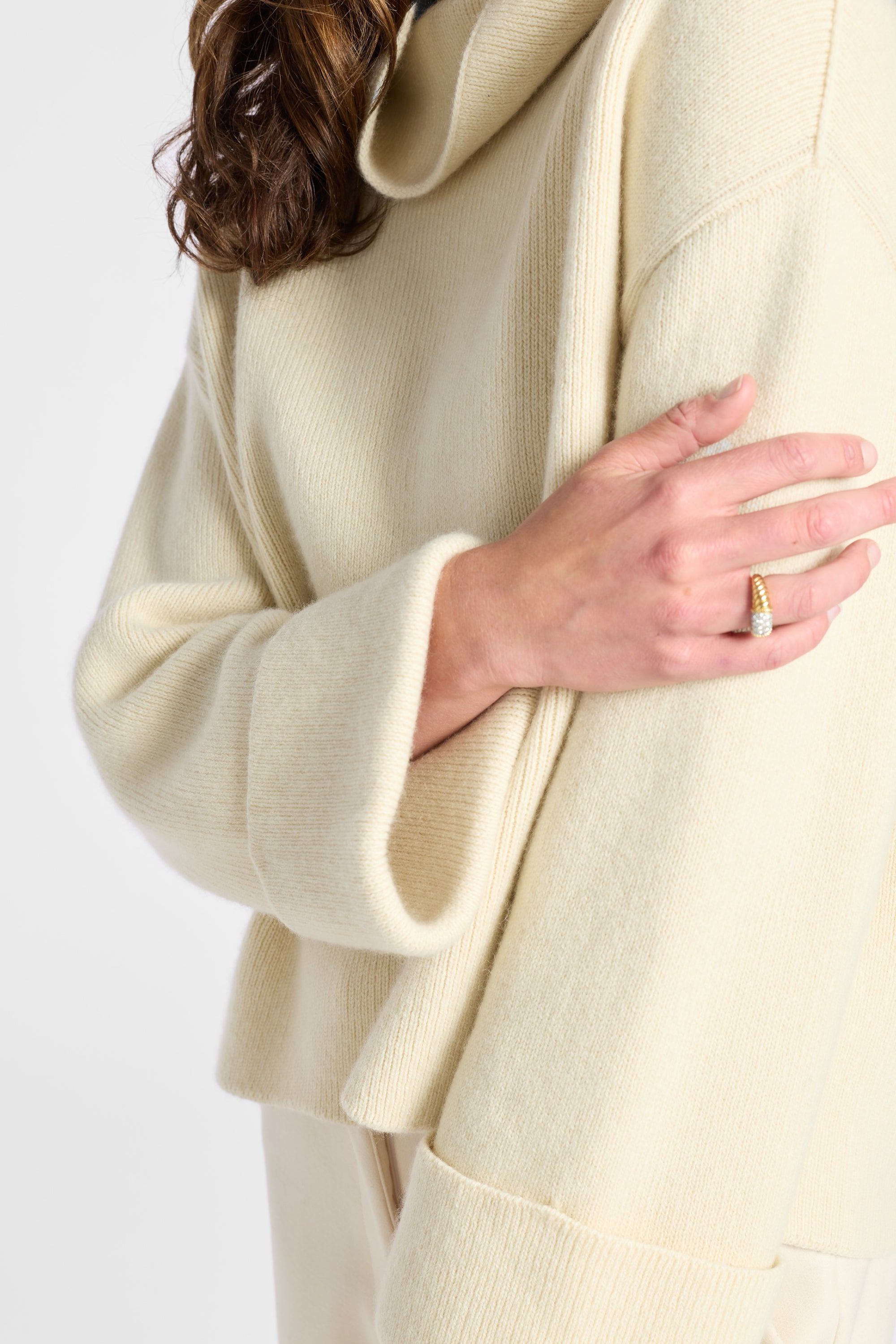 Close-up of a person wearing a cream-colored ribbed knit sweater with a high folded collar and wide cuffs, paired with matching cream pants against a light gray background.