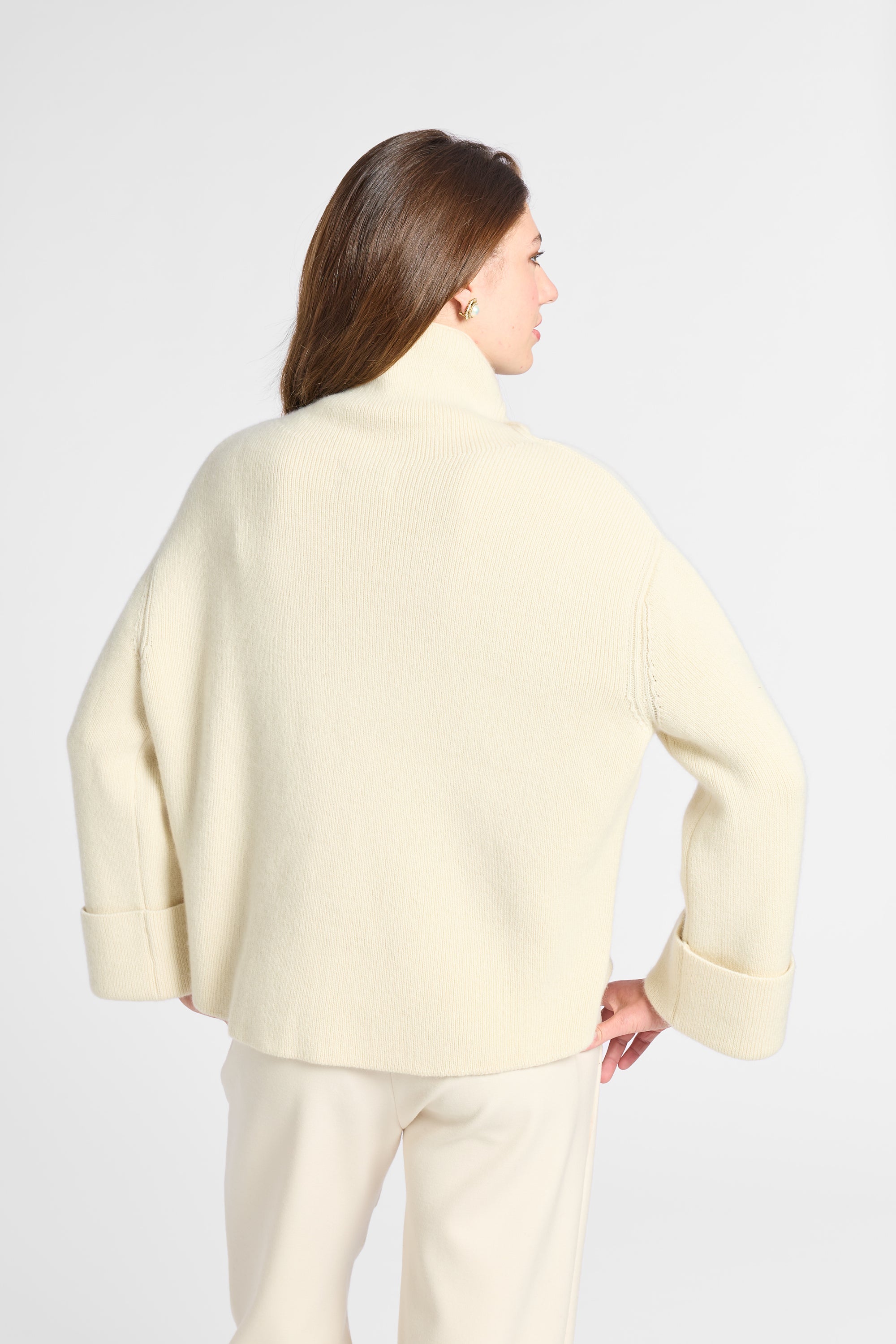 A woman shown from the back wearing a cream-colored knit turtleneck sweater and matching cream trousers against a light gray studio background.