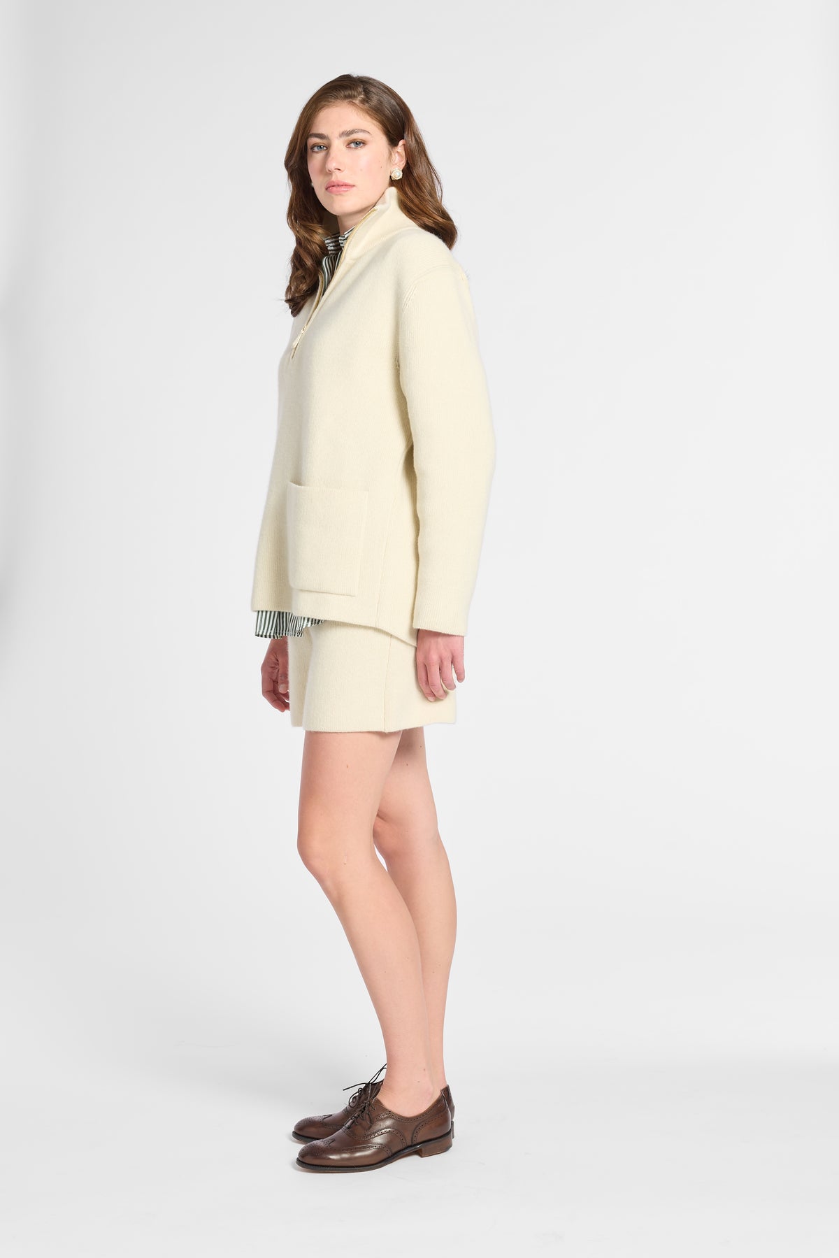 A woman modeling a cream-colored knit sweater and matching mini skirt set, paired with brown leather oxford shoes, against a plain light gray studio background.
