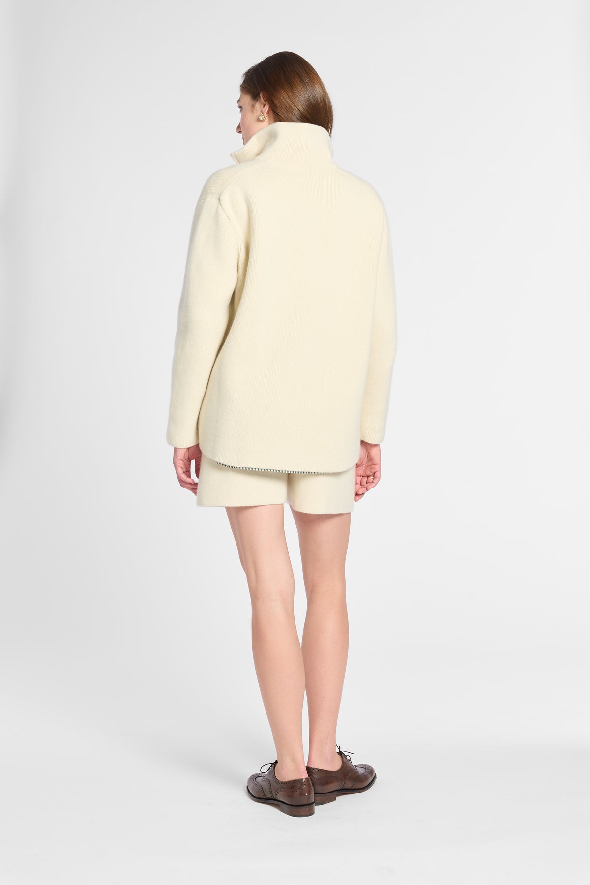 A woman shown from the back wearing a cream-colored knit turtleneck sweater and matching shorts, paired with dark brown leather shoes, against a light gray studio background.