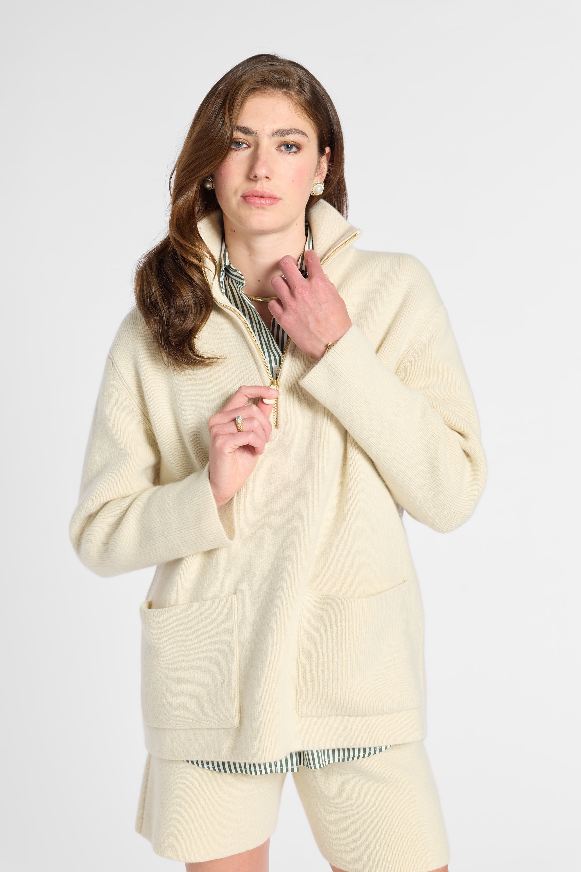 A woman modeling a cream-colored knit zip-up jacket with a high collar and front pockets, paired with matching knit shorts and a striped shirt underneath, against a plain light gray studio background.