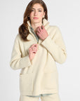 A woman modeling a cream-colored knit zip-up jacket with a high collar and front pockets, paired with matching knit shorts and a striped shirt underneath, against a plain light gray studio background.