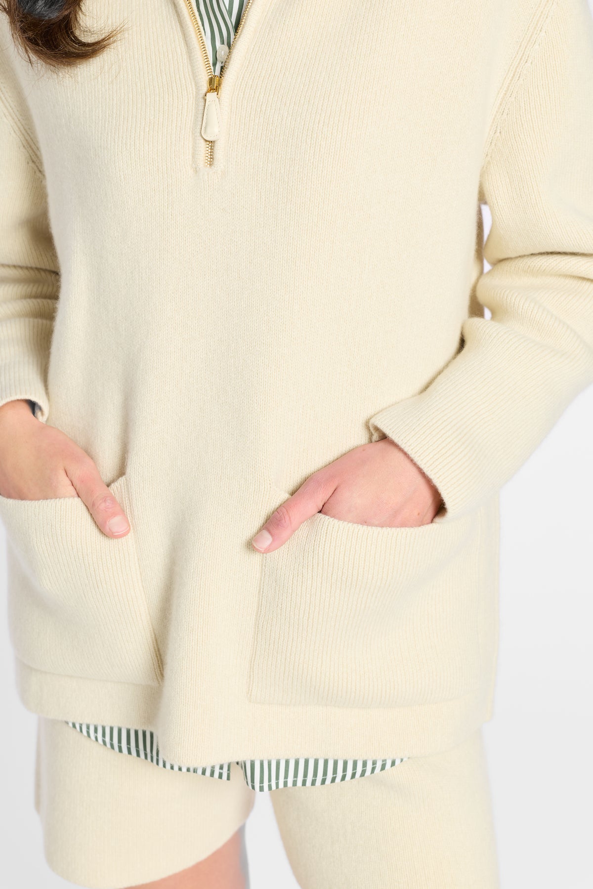 A close-up of a person wearing a cream ribbed knit quarter-zip sweater with front patch pockets, paired with matching knit shorts and a striped shirt peeking out at the hem.