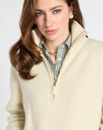 A close-up of a woman wearing a cream-colored ribbed knit quarter-zip sweater over a green and white striped button-down shirt, set against a plain light gray background.