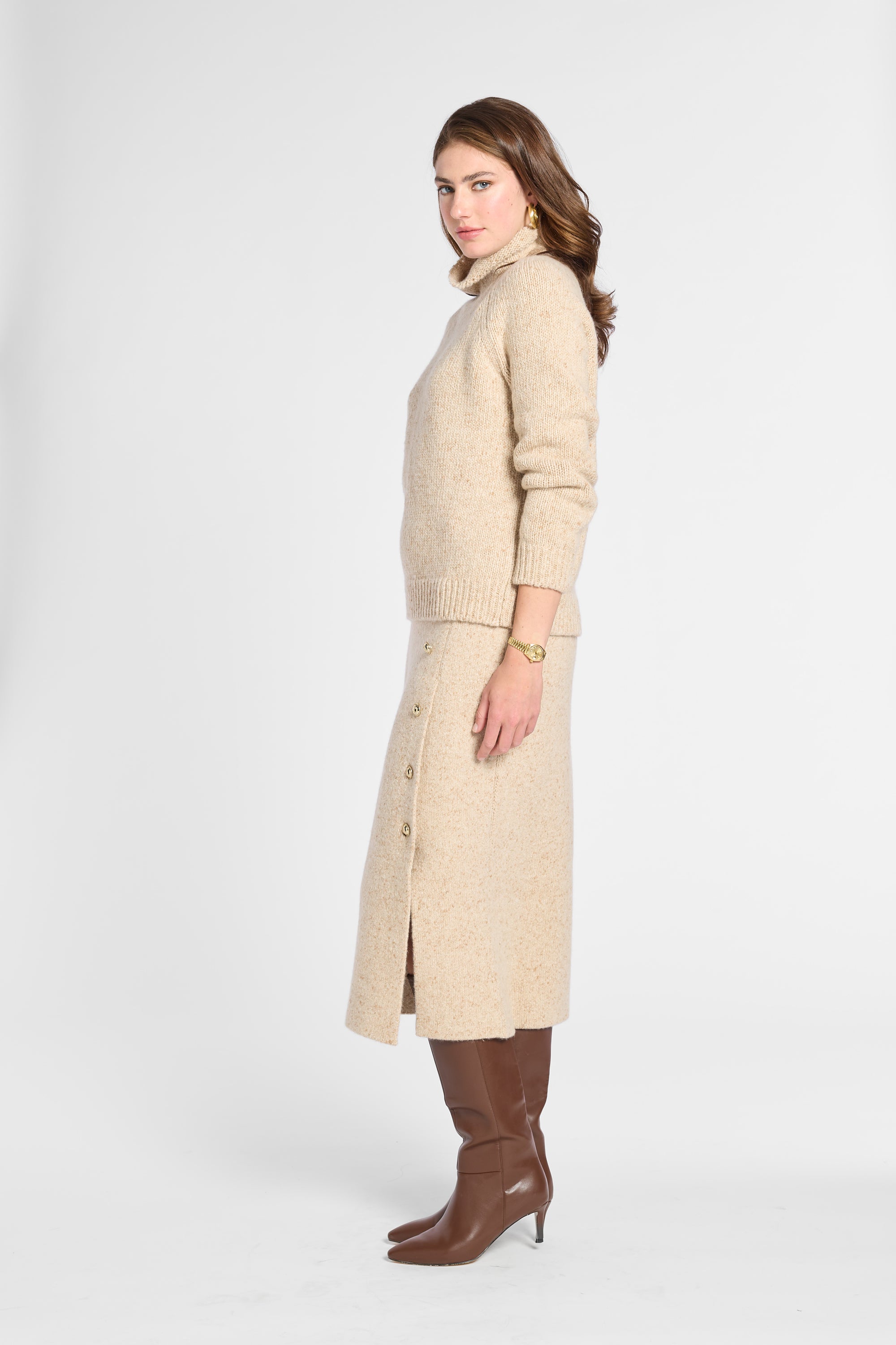 A woman modeling a beige knit turtleneck sweater and matching midi skirt set, paired with brown knee-high boots, against a plain light gray studio background.