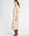 A woman modeling a beige knit turtleneck sweater and matching midi skirt set, paired with brown knee-high boots, against a plain light gray studio background.