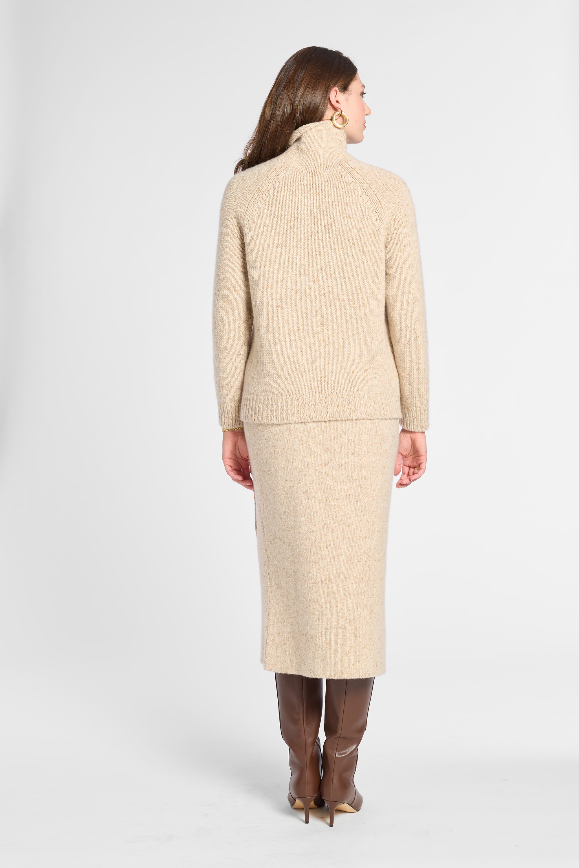 A woman shown from the back wearing a beige knit turtleneck sweater and matching midi skirt, paired with brown knee-high boots, against a light gray studio background.