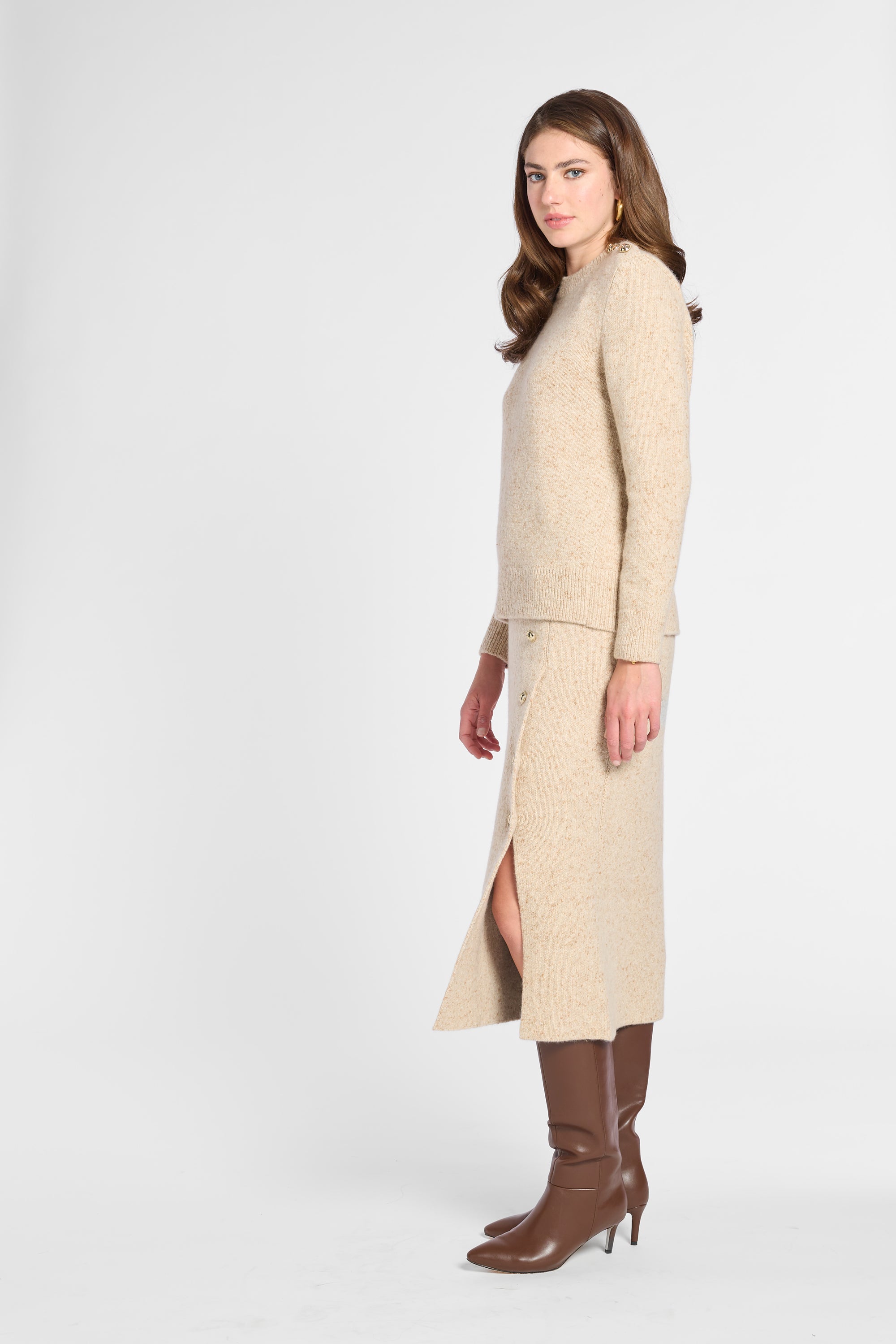 A woman modeling a beige knit sweater and matching midi skirt set with a side slit, paired with brown knee-high heeled boots, against a light gray studio background.