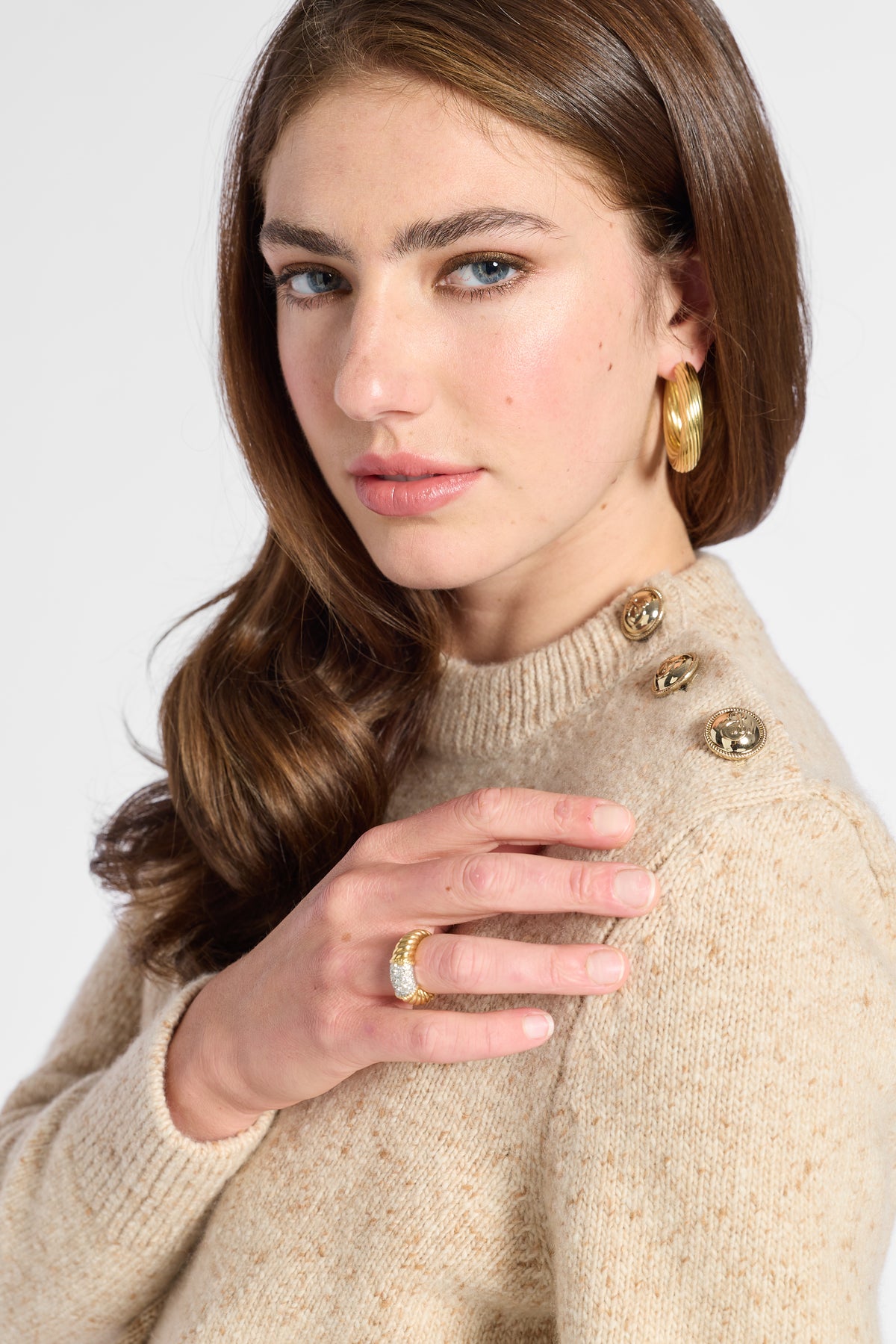 A close-up of a woman wearing a beige knit sweater with decorative gold buttons on the shoulder, a gold ring, and a gold hoop earring, against a light gray studio background.