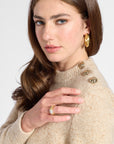 A close-up of a woman wearing a beige knit sweater with decorative gold buttons on the shoulder, a gold ring, and a gold hoop earring, against a light gray studio background.
