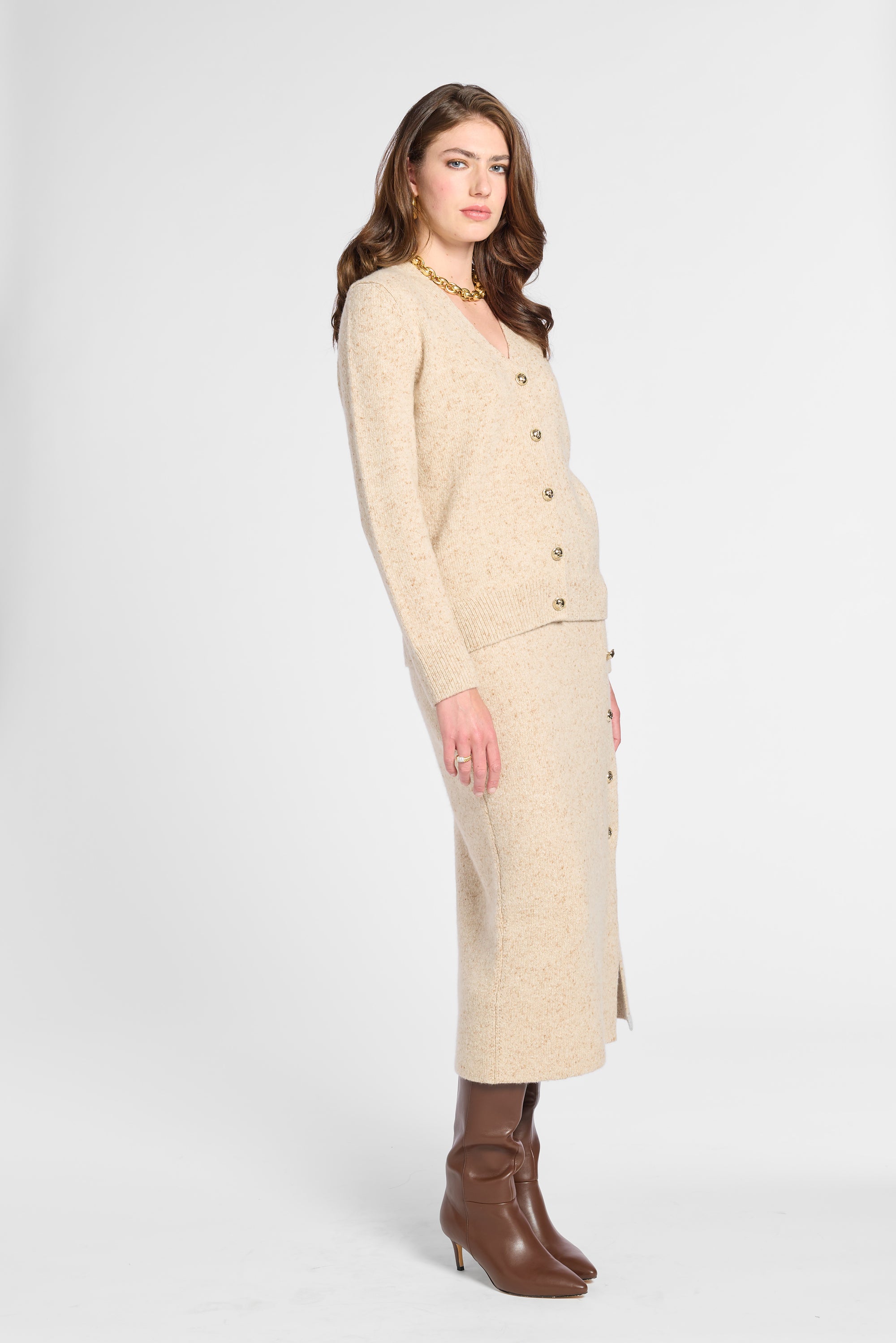 A woman models a beige knit cardigan and matching midi skirt set with gold buttons, paired with brown leather ankle boots, against a light gray studio background.