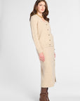 A woman models a beige knit cardigan and matching midi skirt set with gold buttons, paired with brown leather ankle boots, against a light gray studio background.