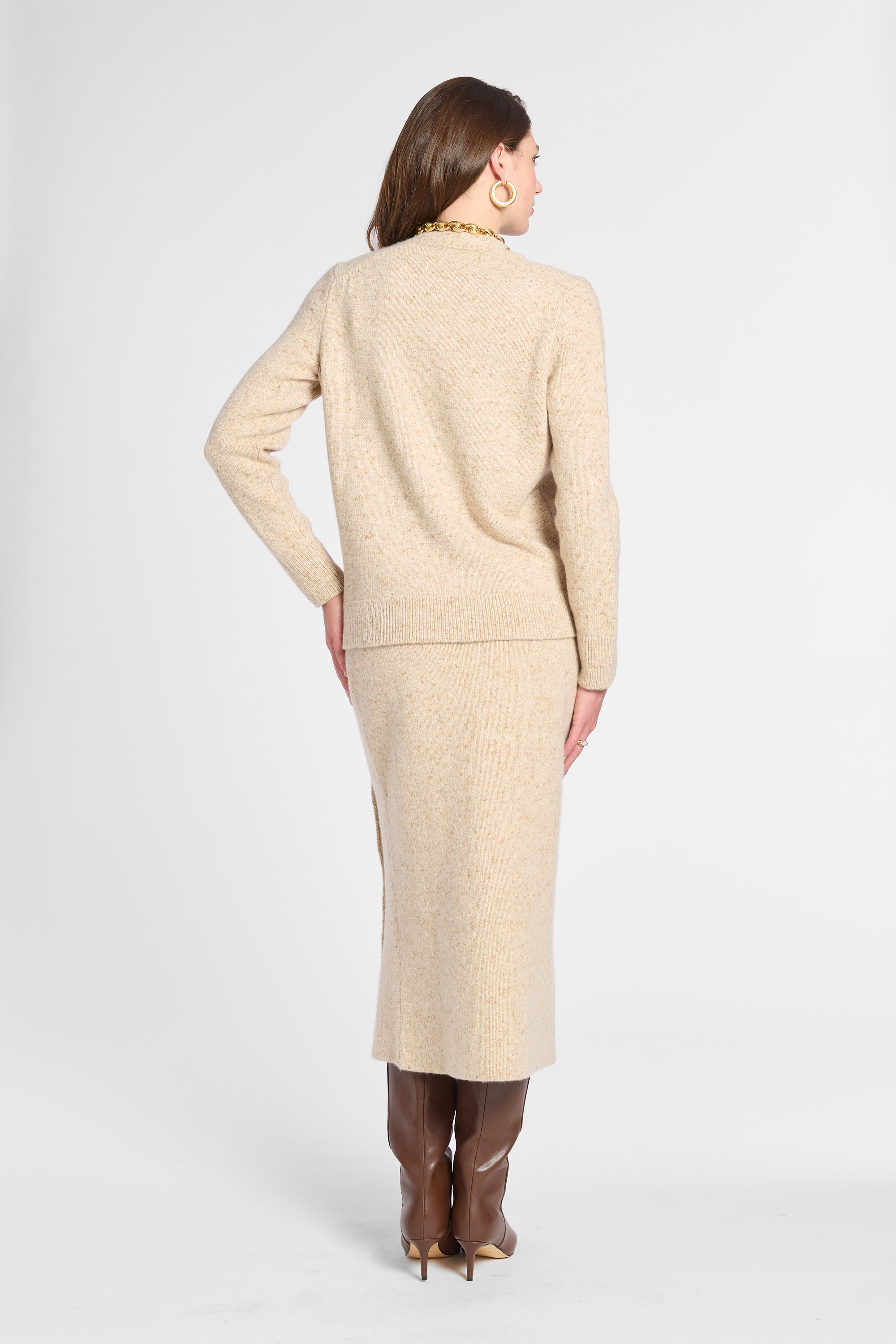 A woman shown from the back wearing a light beige knit sweater and matching midi skirt set, paired with brown knee-high boots, against a plain light gray studio background.