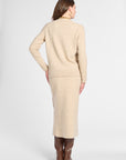 A woman shown from the back wearing a light beige knit sweater and matching midi skirt set, paired with brown knee-high boots, against a plain light gray studio background.