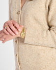 Close-up of a person wearing a beige knit cardigan with gold buttons and a gold watch on the wrist, against a clean white background.