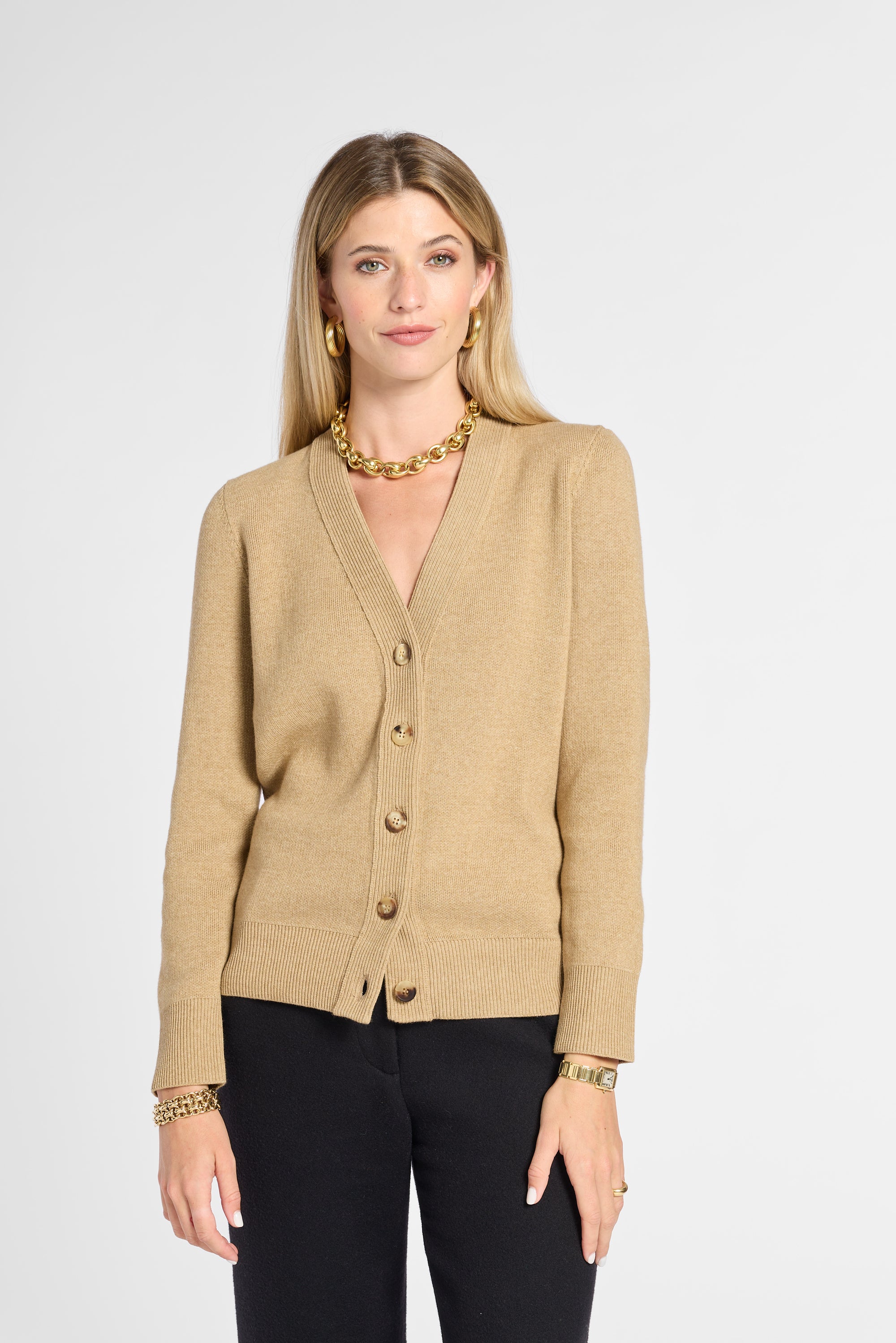A woman modeling a beige knit cardigan with gold buttons, paired with black pants and gold jewelry, against a light gray studio background.