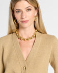 A woman with blonde hair wearing a chunky gold chain necklace, matching gold hoop earrings, and a beige knit cardigan against a light gray background.