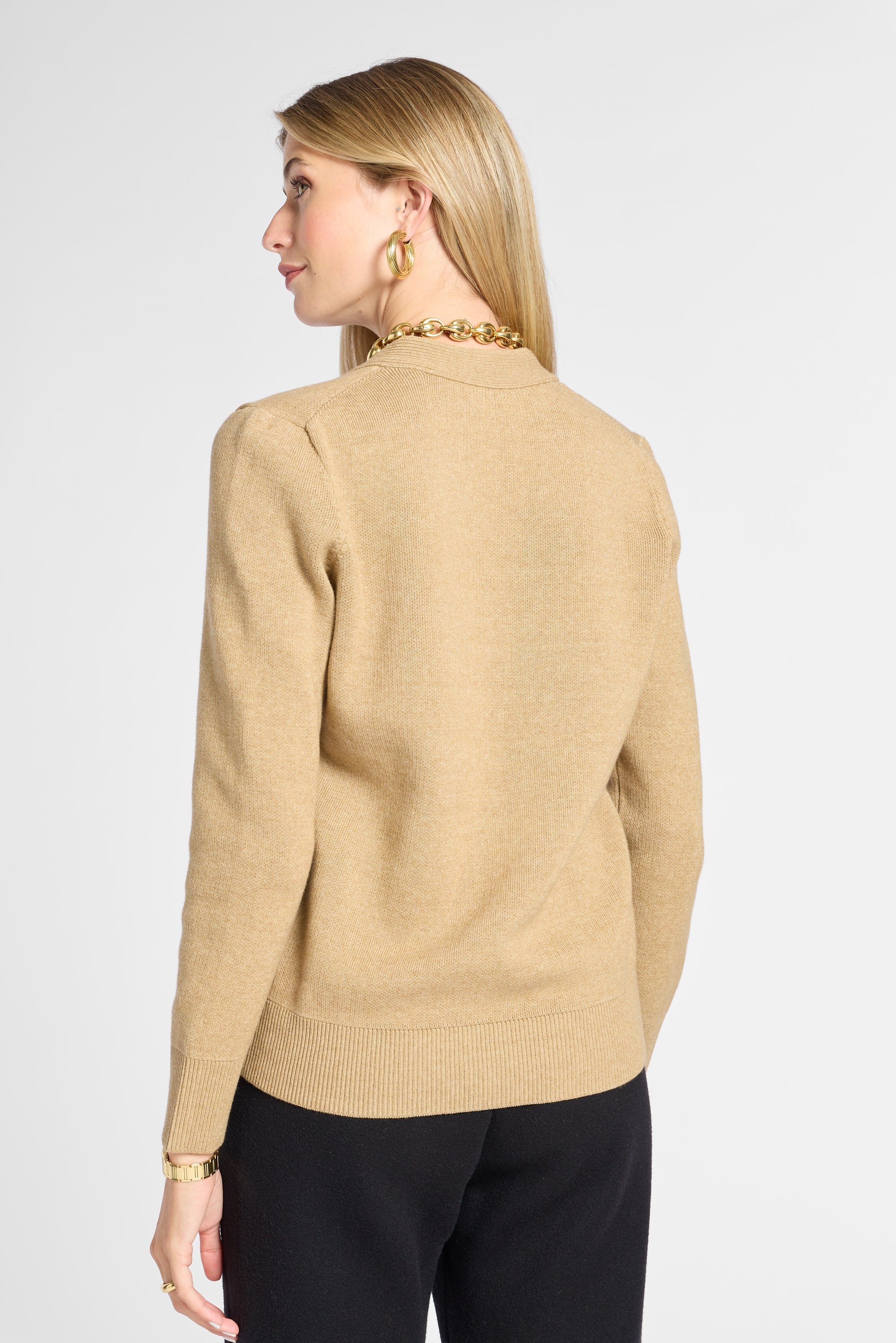 A woman shown from the back wearing a beige knit sweater with a gold chain detail around the collar, paired with black pants, against a light gray studio background.