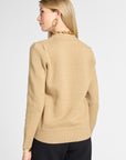 A woman shown from the back wearing a beige knit sweater with a gold chain detail around the collar, paired with black pants, against a light gray studio background.