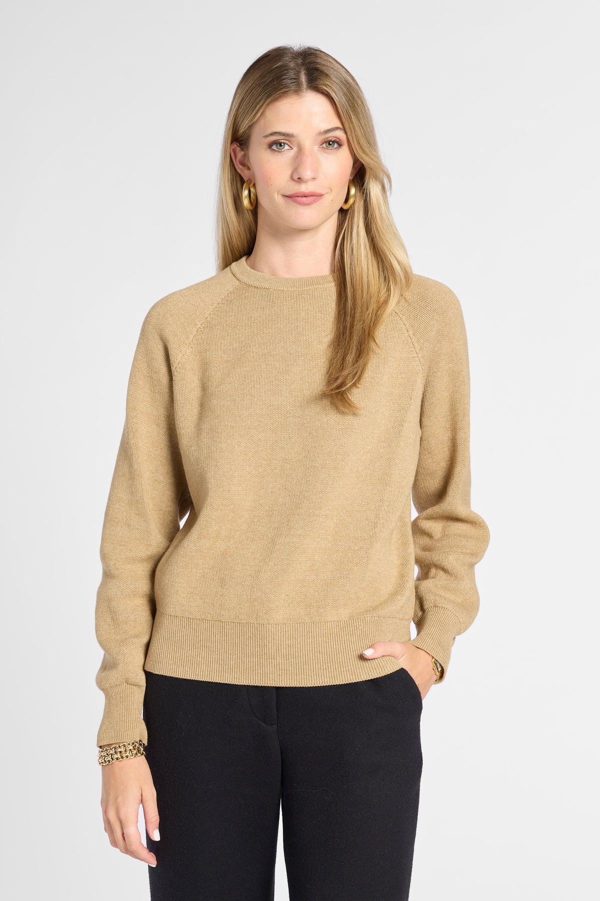 A woman modeling a beige crewneck knit sweater with black pants, standing against a light gray studio background.