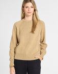 A woman modeling a beige crewneck knit sweater with black pants, standing against a light gray studio background.