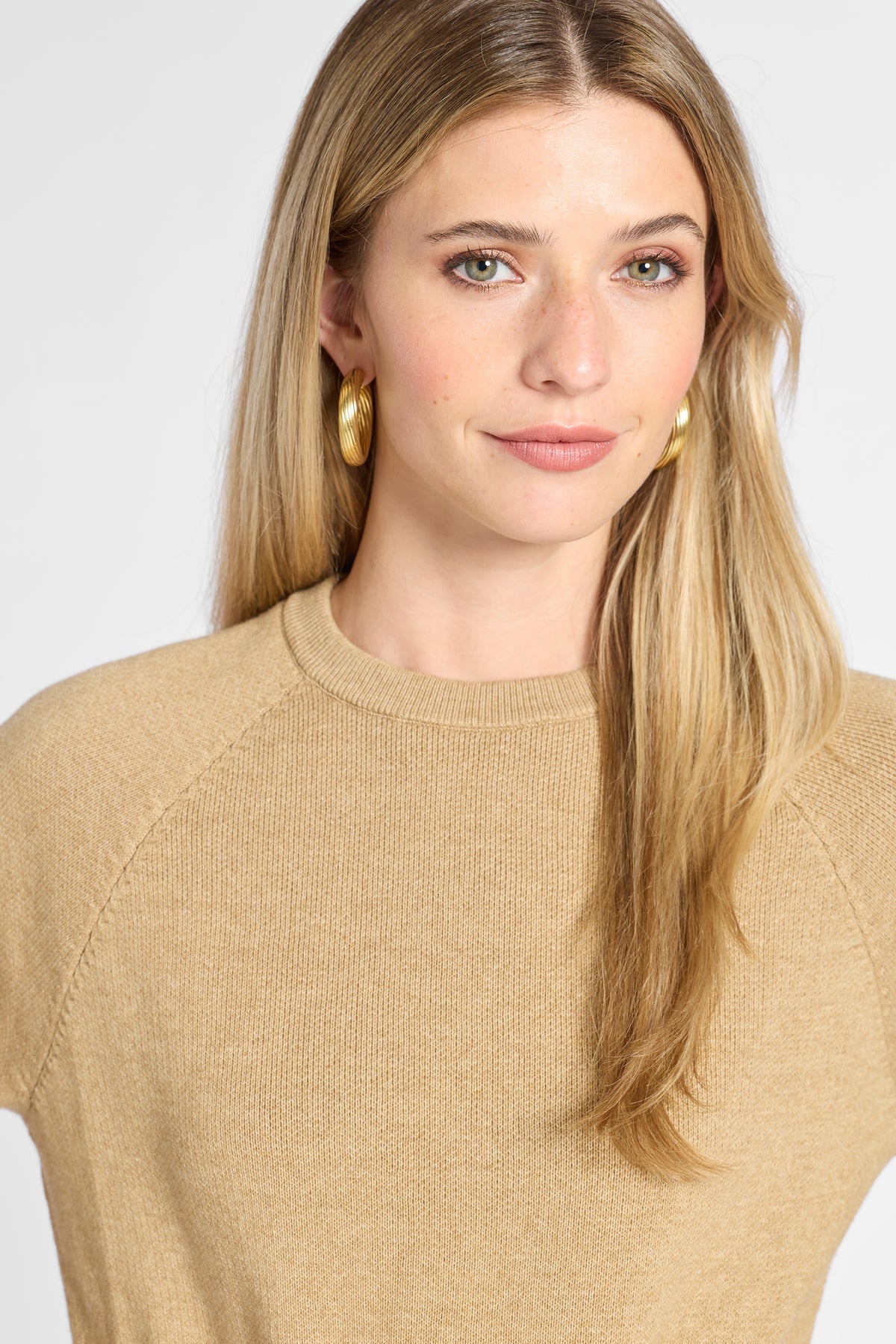 A close-up of a woman with blonde hair wearing a beige knit crewneck sweater and gold hoop earrings against a light gray background.