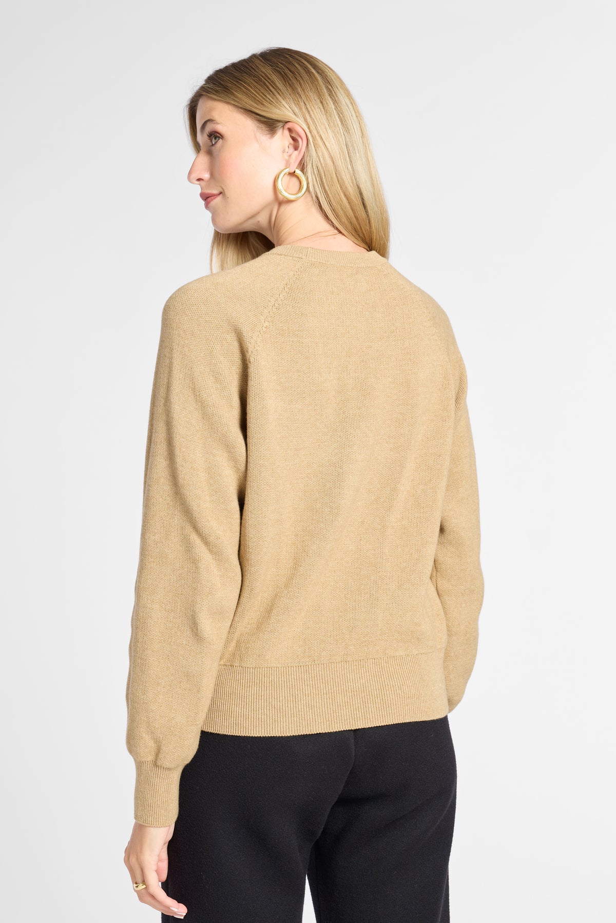 A woman shown from the back wearing a light tan knit sweater and black pants against a plain light gray studio background.