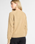 A woman shown from the back wearing a light tan knit sweater and black pants against a plain light gray studio background.