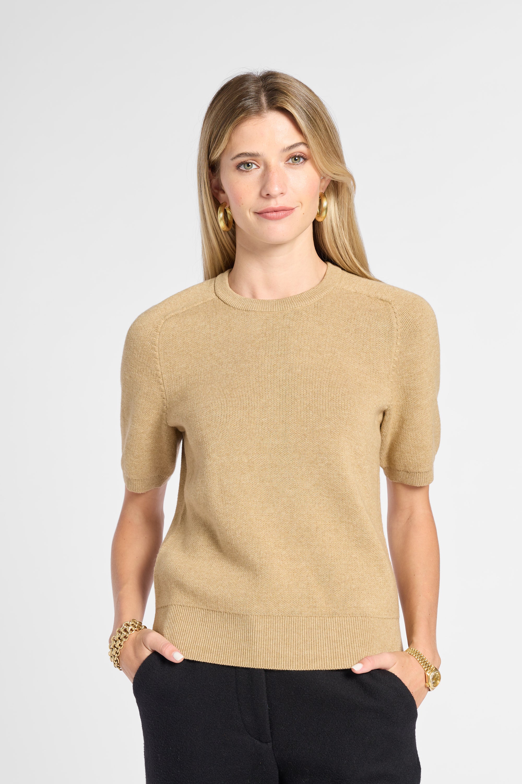 A woman modeling a short-sleeve knit sweater in a warm beige color, paired with black pants, against a light gray studio background.
