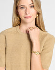 A woman wearing a short-sleeve crewneck knit sweater in a warm tan/beige color, styled with gold hoop earrings, a gold watch, and a gold ring against a light gray background.
