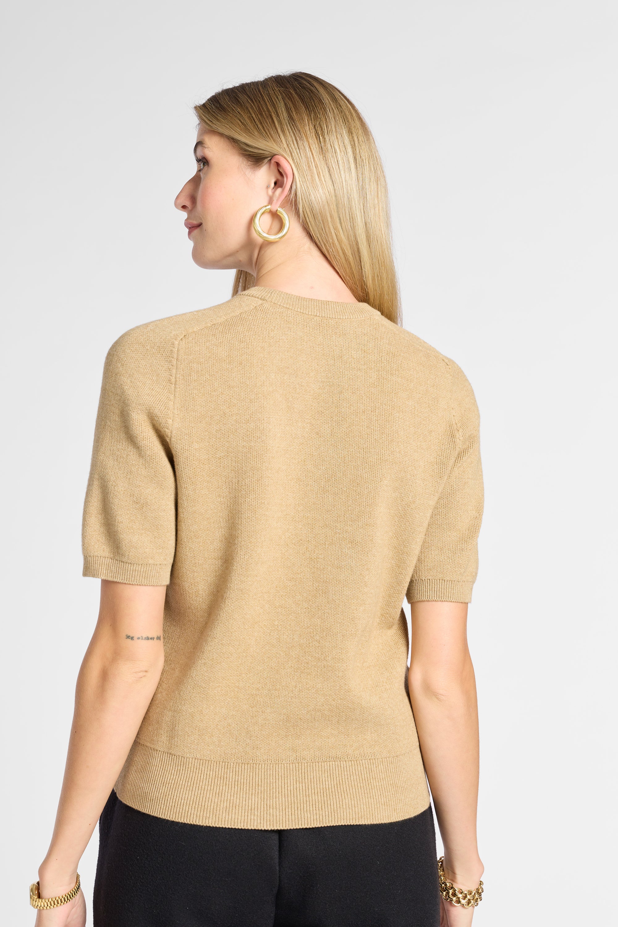 A woman shown from the back wearing a beige short-sleeve knit top and black pants against a light gray studio background.