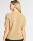 A woman shown from the back wearing a beige short-sleeve knit top and black pants against a light gray studio background.