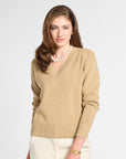 A woman modeling a beige V-neck knit sweater with cream pants, a pearl necklace, and a gold watch against a light gray studio background.