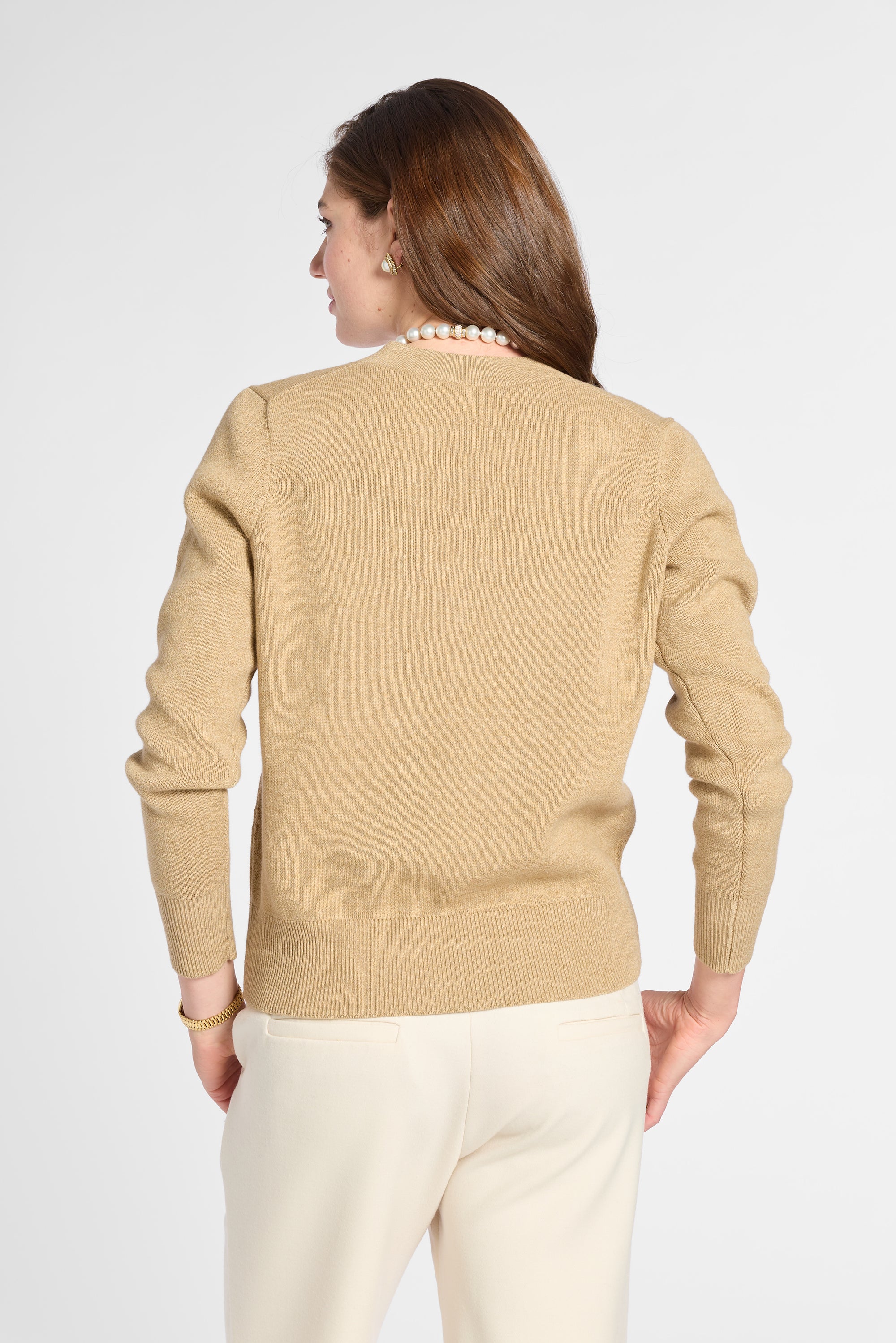 A woman shown from the back wearing a beige knit sweater and cream pants against a light gray studio background.