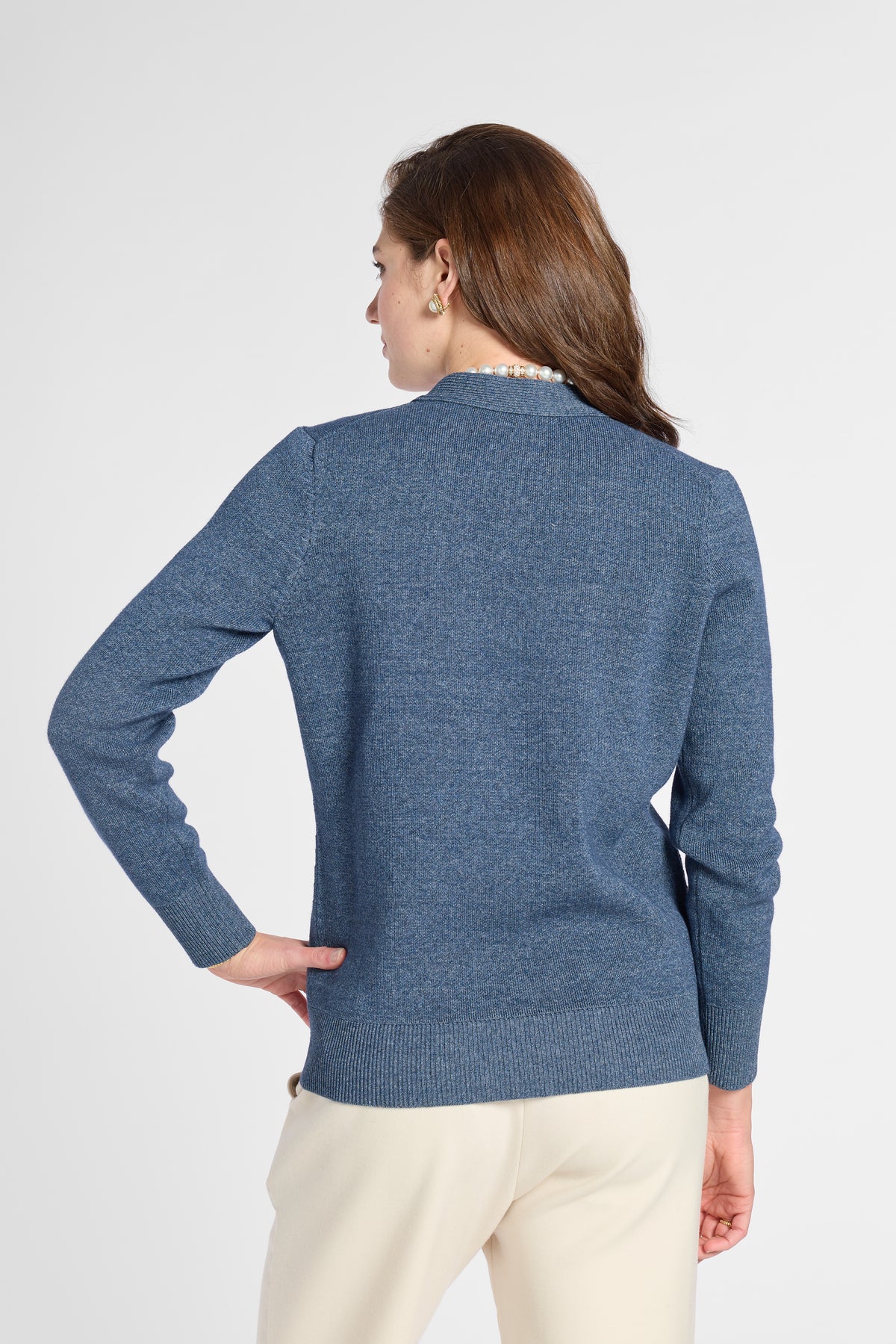 A woman shown from the back wearing a heathered blue long-sleeve knit sweater and cream pants against a light gray studio background.